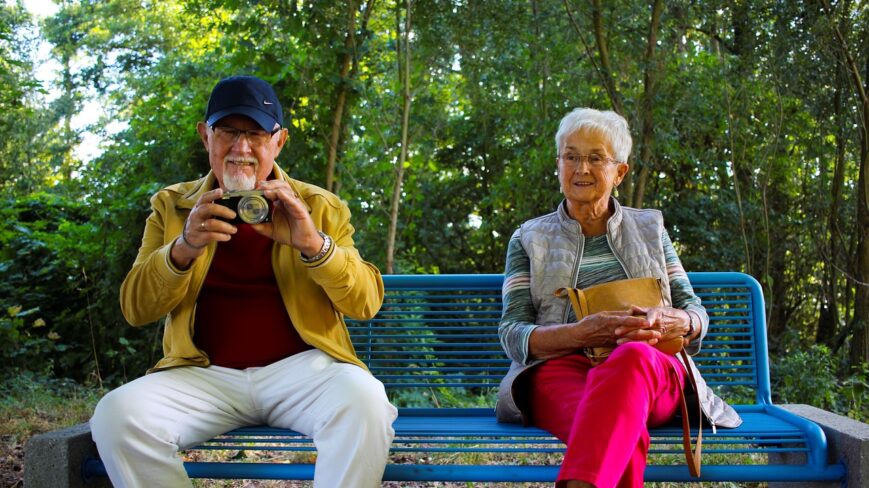 Kaksi senioria istuu puistonpenkillä, mies kuvaa kameralla ja nainen katsoo puistoon päin.