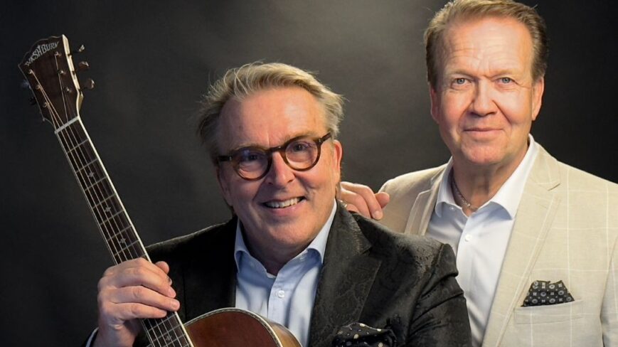 Kuvassa iloisesti hymyilevät viihdetaiteilijat Heikki Hilander sekä Joel Hallikainen. Joel Hallikaisella on sylissään kitara.