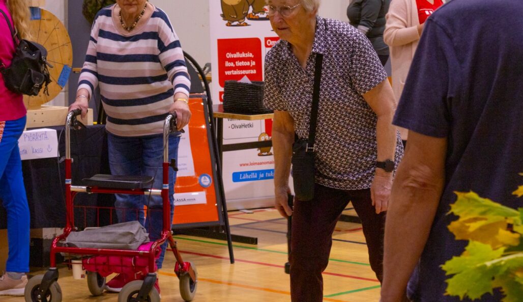 Kaksi iäkästä naista pelaamassa jotain peliä, mies katsoo vieressä, iloisessa senioripäivän tapahtumassa.