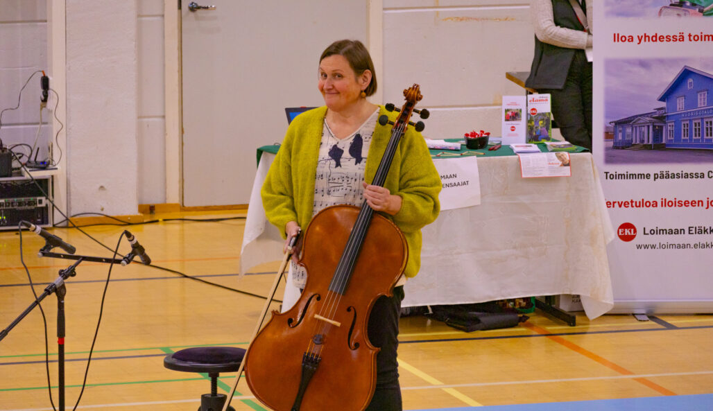 Nainen seisoo salissa hänellä on sellosoitin edessään ja jousi toisessa kädessä.