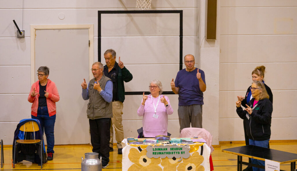 Ikäihmisiä, miehiä ja naisia seisomassa liikuntasalissa. He tekevät yhdessä sormijumppaa.