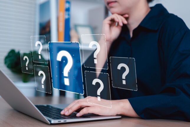 Kuvassa henkilö naputtaa yhdellä kädellä tietokonetta ja ympärillä leijuu kysymysmerkkejä