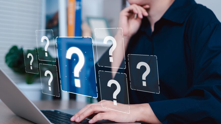 Kuvassa henkilö naputtaa yhdellä kädellä tietokonetta ja ympärillä leijuu kysymysmerkkejä