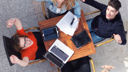 Neljä iloista nuorta istuu pöydän ääressä, katsoo ylöspäin ja hurraa. Pöydällä on tietokone ja muistiinpanovälineitä.