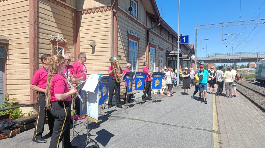 Loimaan soittokunnan 7 jäsentä soittavat puhaltimia ja heidän takanaan on Loimaan rautatieasema.